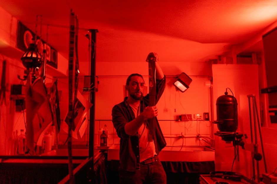 A photographer in a red-lit darkroom carefully examines a film strip during the developing process.