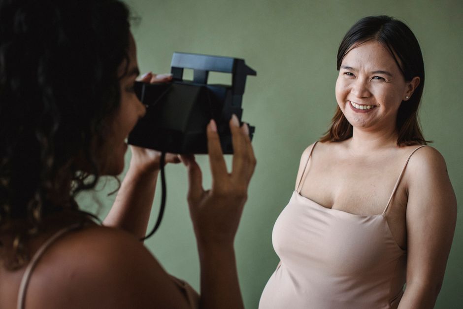 Two women in a studio setting, one photographing the other, both smiling.
