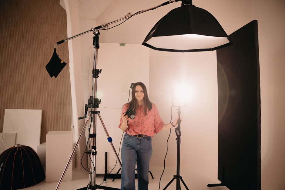 Female photographer holding camera in a well-lit studio with professional lighting and equipment.