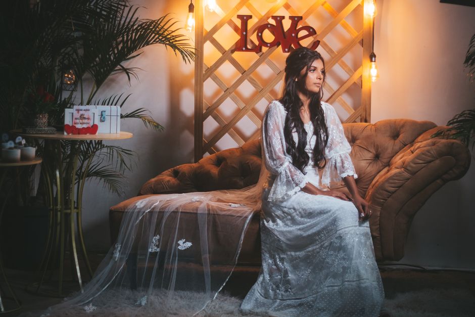 Bride in a lace wedding dress sitting on a vintage couch in a cozy, boho style setting with soft lighting.